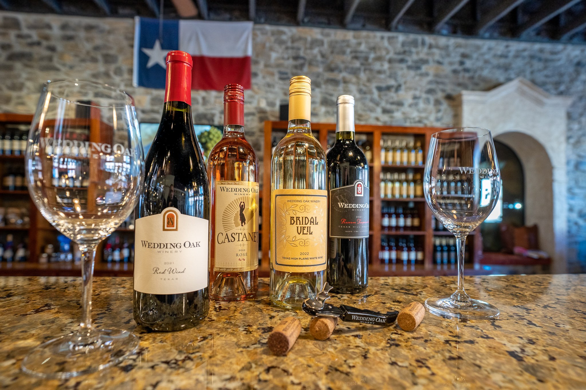Four bottles of Wedding Oak wine and two glasses on the tasting-room bar, a Texas flag on the stone wall behind