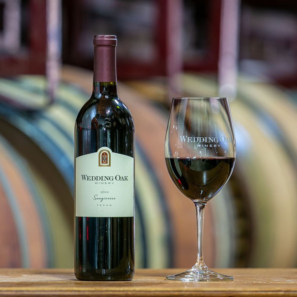 A bottle of Sangiovese beside a glass of red, barrels behind
